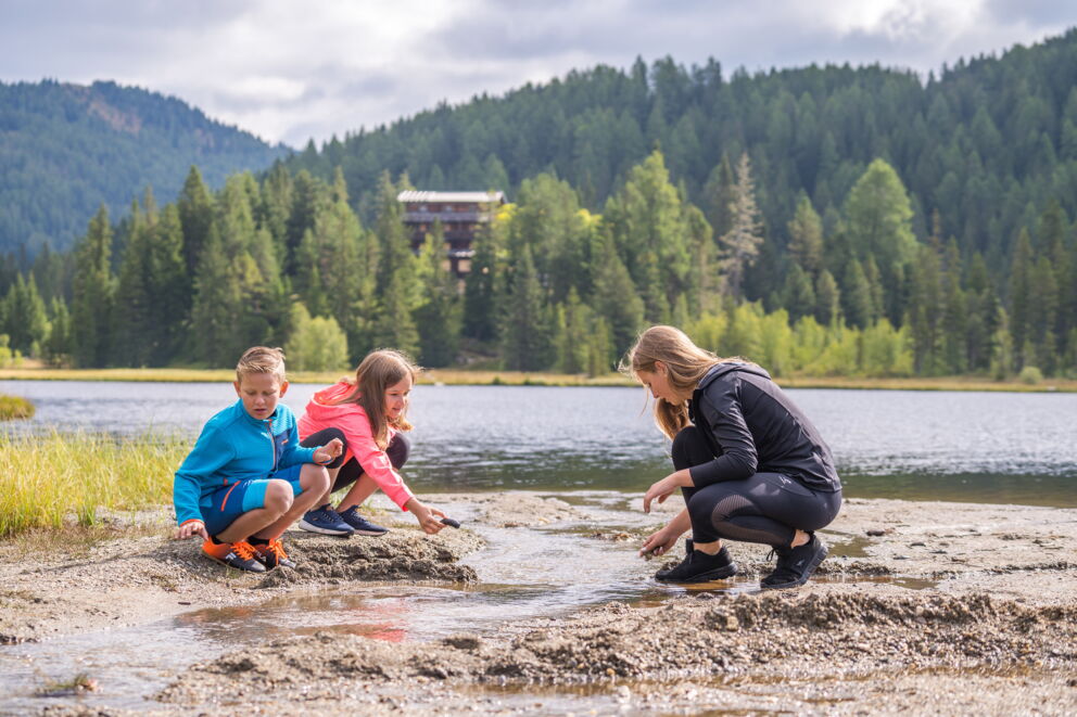 Family playing on the lakeshore with forest and mountains in the background.