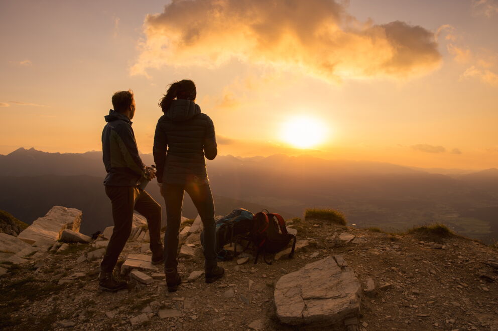 Two people enjoy the sunset on a mountain peak with a wide view of the valley.