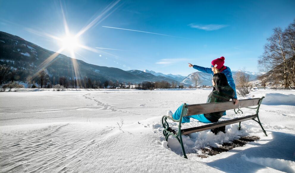 Zwei Frauen sitzen auf einer Bank im Freien im Schnee und genießen die winterliche Landschaft, eine Frau zeigt auf die strahlende Sonne.