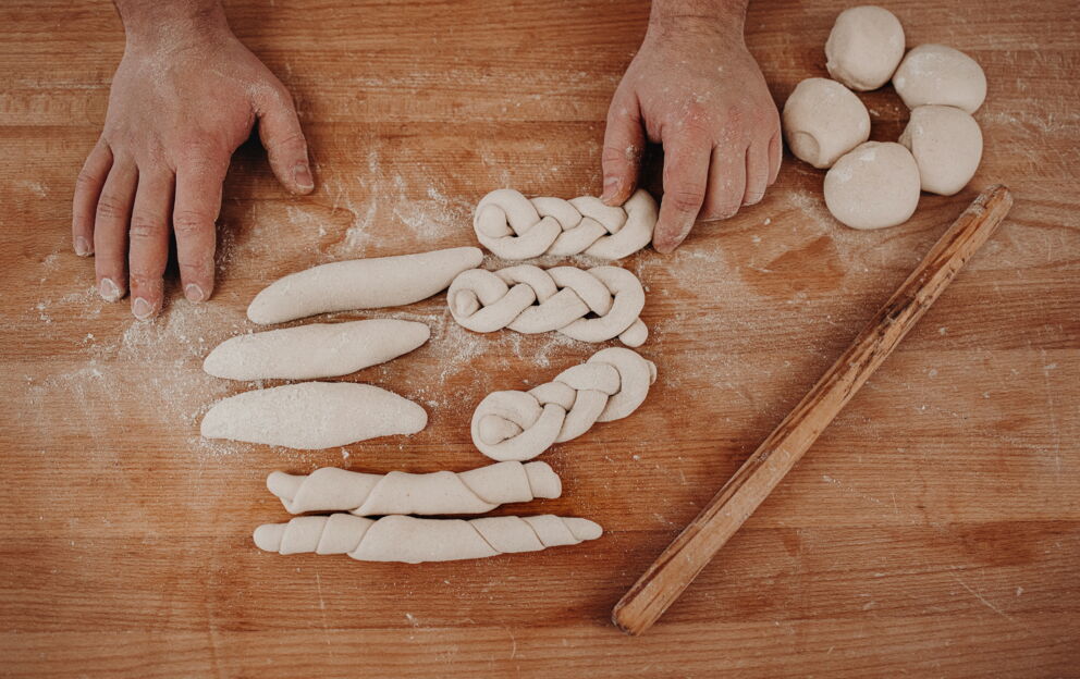 Hände formen Teigstücke auf bemehlter Holzfläche mit einem Nudelholz.