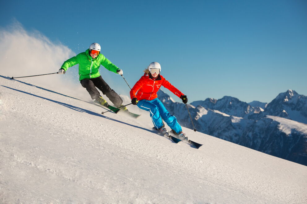 Zwei Skifahrer in bunter Kleidung fahren gemeinsam eine schneebedeckte Piste hinunter.