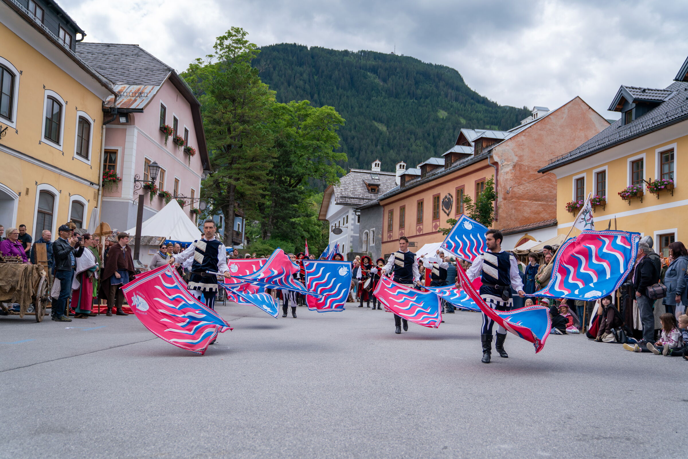 Teilnehmer in traditionellen Kostümen führen bunte Flaggen in belebter Dorfstraße.