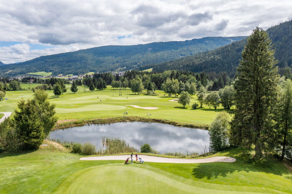 Golfer auf gepflegtem Grün mit malerischem Teich und Wald im Hintergrund.