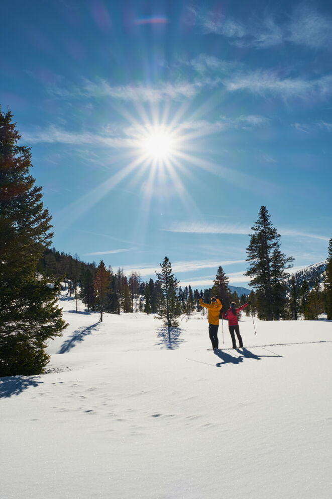 Zwei Personen genießen den sonnigen Wintertag auf verschneitem Gelände mit klarblauem Himmel.