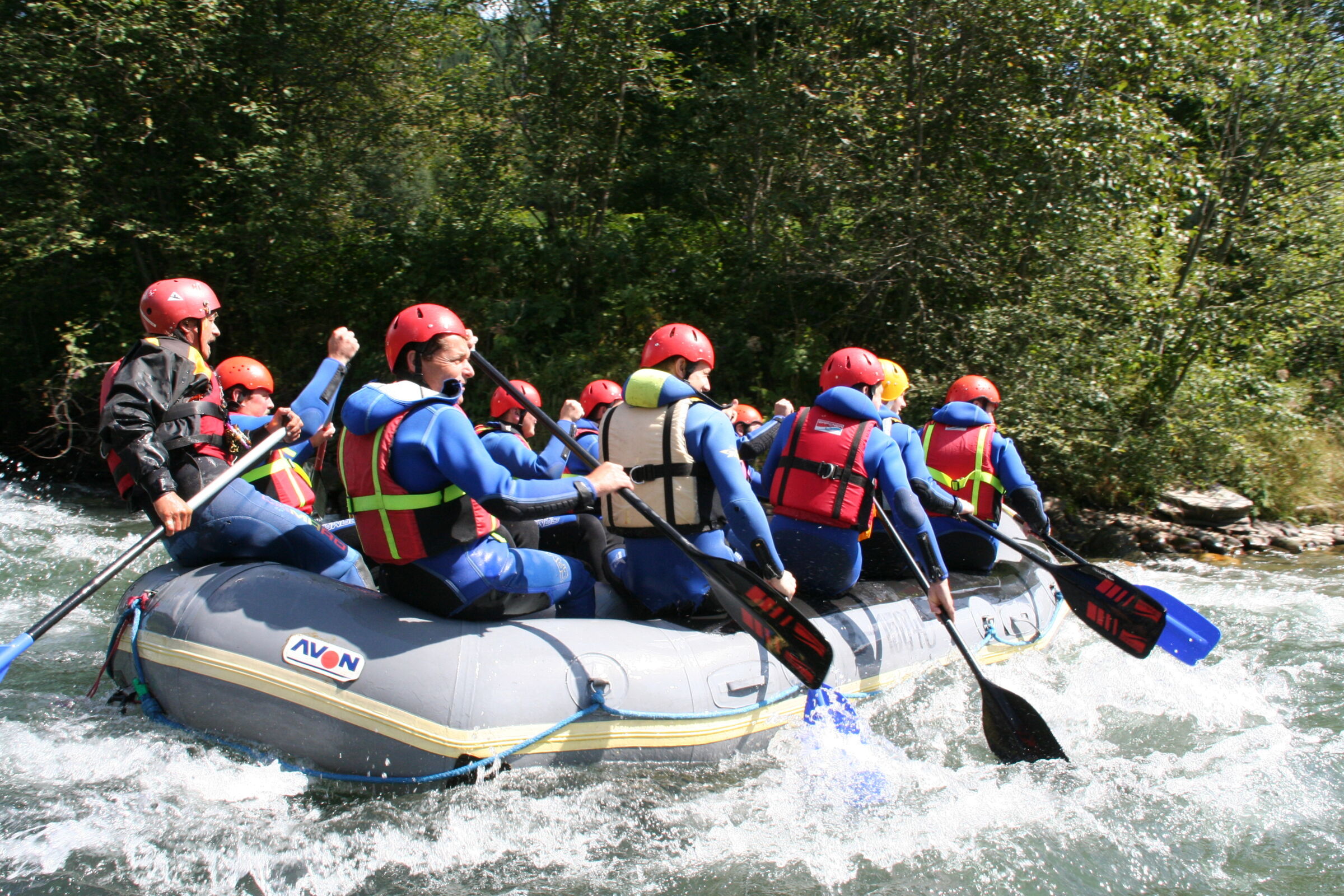 Gruppe von Personen beim Rafting auf einem Fluss mit Schutzhelmen und Schwimmwesten.