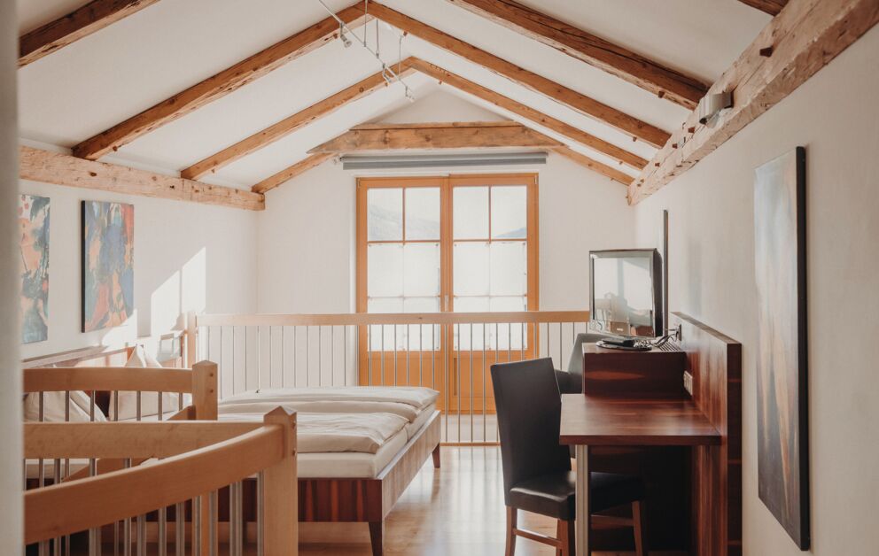 Helles Dachzimmer mit Holzbalken, Schreibtisch und großem Fenster für natürlichen Lichteinfall.