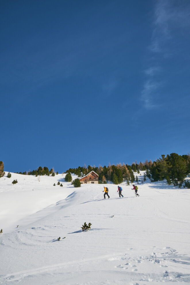 Drei Wanderer auf verschneitem Bergpfad vor rustikaler Berghütte unter strahlend blauem Himmel.