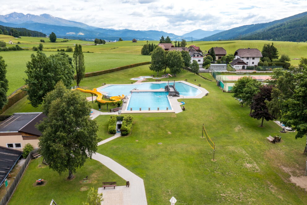 Weitläufige Poollandschaft mit grüner Wiese und Wasserrutsche, umgeben von Bergen.