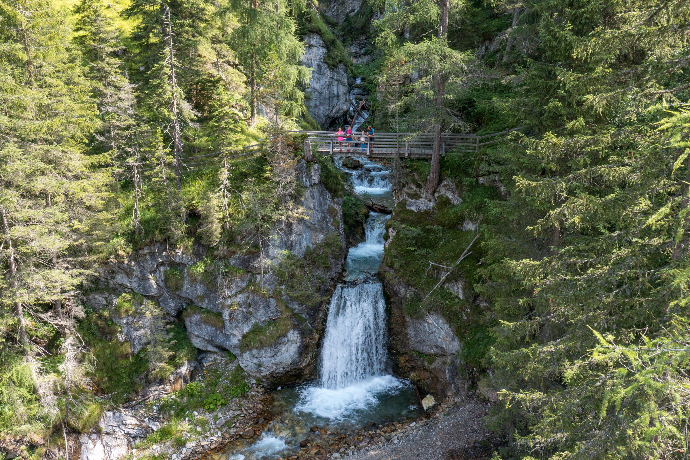 Ein ruhiger Wasserfall fließt durch einen dichten, grünen Wald mit hölzernem Steg.