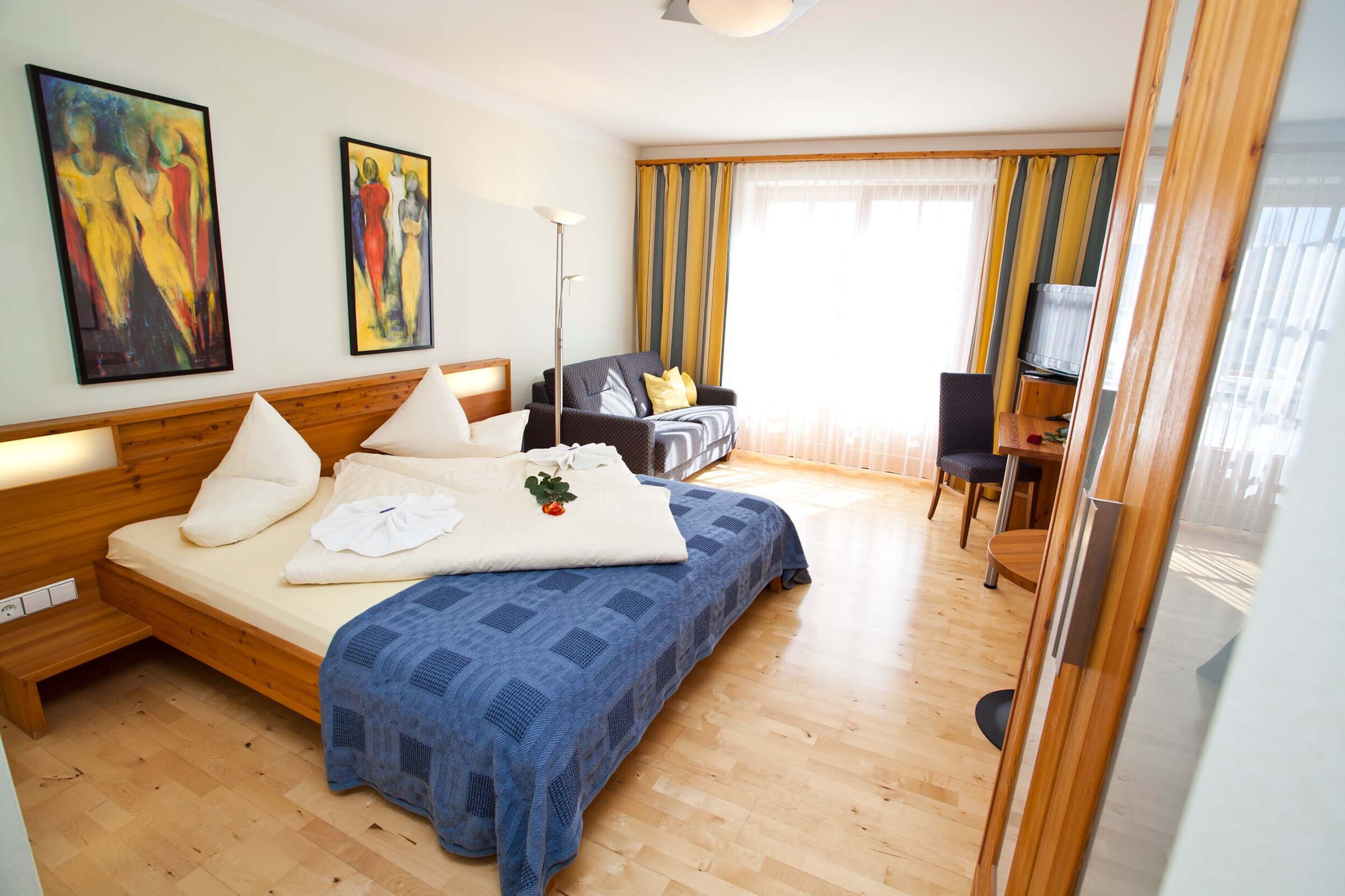Helles Zimmer mit Holzmöbeln, Sofa, Kunstwerken und großem Fenster mit Vorhängen.